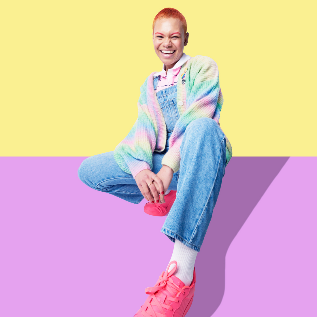 A smiling individual with short-cropped red hair wearing a pastel rainbow cardigan and denim overalls crouches against a split yellow and purple background.