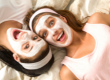 Two friends lie side-by-side on light-colored bedding, both wearing white facial masks and smiling enthusiastically.
